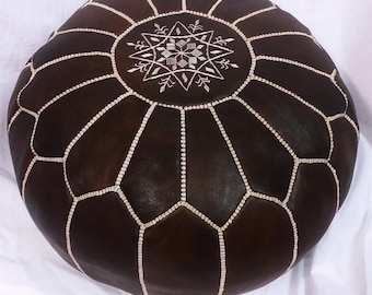 May include: A round, dark brown leather pouf with white stitching and a central starburst design. The pouf is a decorative piece of furniture, likely used as a footrest or extra seating. The stitching creates a geometric pattern.