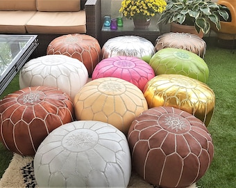 May include: A collection of colorful Moroccan leather poufs in various shades, including brown, white, pink, and gold. The round ottomans are arranged on a green surface, suggesting outdoor or patio decor.