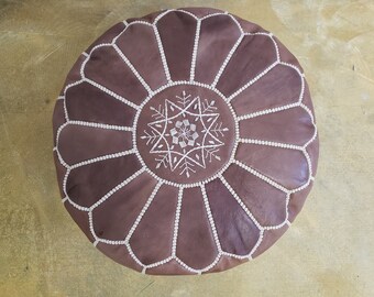 May include: Round brown leather ottoman with a decorative white embroidered starburst design in the center. The ottoman is divided into sections and stitched with white thread. A stylish piece of furniture.