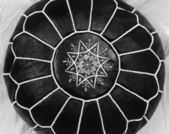May include: Black round leather ottoman with white stitching and a central starburst design. The ottoman features a geometric pattern and is a decorative piece for home decor.