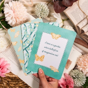 Könnte beinhalten: Eine Hand hält eine türkisfarbene Karte mit einem Zitat über Bücher und Schmetterlinge, umgeben von anderen Karten mit Schmetterlings- und Blumenmustern. Die Szene umfasst offene Bücher und dekorative Blumen in Rosa und Weiß.