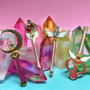 以下が含まれることがあります： セーラームーンのアニメから、魔法の杖の形をした金色のエナメルピンが5つ。杖は虹色のピンク、イエロー、グリーン、ブルーのクリスタルの上に飾られています。