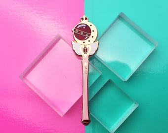 May include: A rose gold-colored wand with a crescent moon and crown design, featuring a red center and white wings. The wand is set against a vibrant pink and teal background, with clear square blocks adding depth.