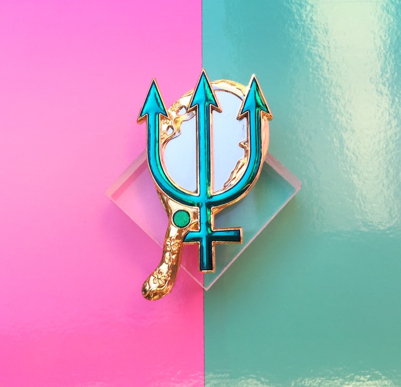 May include: Gold-tone pin with a green trident design and a white mirror-like surface. The pin is on a pink and blue background.