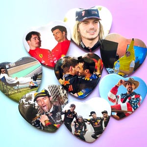 May include: A collection of heart-shaped magnets featuring images of Formula 1 drivers and other related images. The magnets have a glossy finish and are arranged on a gradient pink and blue background.
