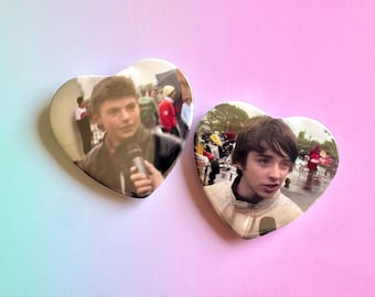May include: Two heart-shaped buttons featuring images of a person holding a microphone. The buttons have a glossy finish and are set against a gradient pink and blue background. The images appear to be from a live event.