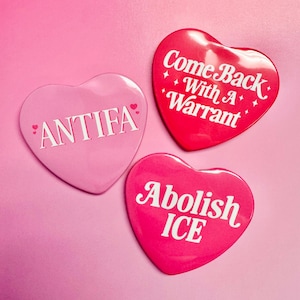 Puede incluir: Tres botones en forma de corazón con texto blanco sobre fondo rosa. Uno dice "ANTIFIA" en rosa, otro "Come Back With A Warrant" en rojo, y el tercero "Abolish ICE" en rosa.