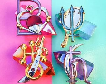 May include: Five colorful enamel pins featuring a heart with a crown, a trident with a mirror, a sword with a circle, a cross with a crescent moon, and a star with a crescent moon. The pins are displayed on iridescent surfaces.