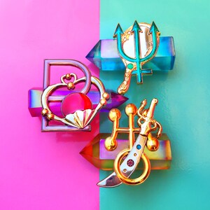 May include: A set of five gold-tone enamel pins featuring a heart, a trident, a sword, and two other abstract designs. The pins are arranged on a pink and blue background.