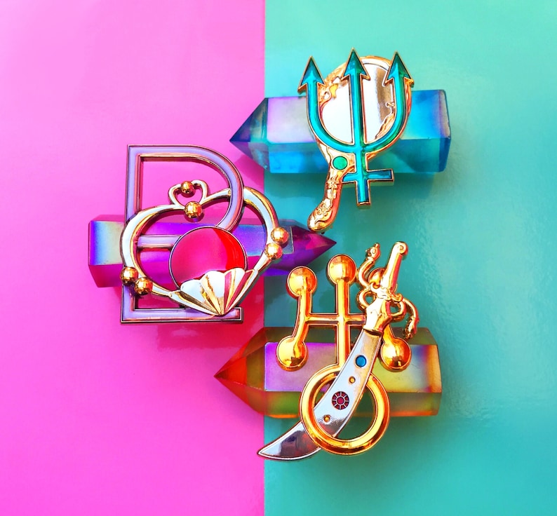 May include: A set of five gold-tone enamel pins featuring a heart, a trident, a sword, and two other abstract designs. The pins are arranged on a pink and blue background.