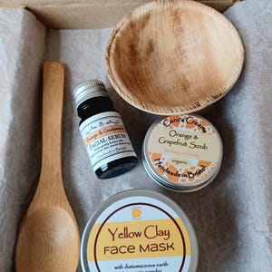 May include: A skincare set containing a wooden spoon, a small palm leaf bowl, a dark glass bottle of Orange & Cardamom Facial Serum, a tin of Orange & Grapefruit Scrub, and a tin of Yellow Clay Face Mask with turmeric powder and diatomaceous earth.