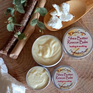May include: A close-up of shea butter products. Two small tins of balm are next to a larger open tin of balm. The tins are labelled "Caro's Creams Shea Butter Rescue Balm". White shea butter chunks and a wooden spoon are also visible.