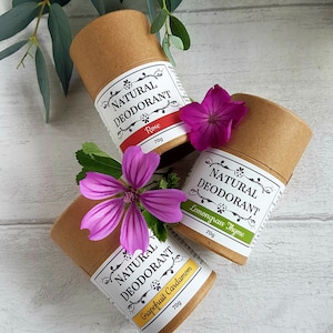 May include: Three cylindrical cardboard tubes of natural deodorant, each weighing 70g. The labels feature the text Natural Deodorant with scent names Rose, Lemongrass Thyme, and Grapefruit Cardamom. The tubes are decorated with purple flowers.