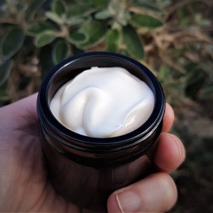 May include: A small, cylindrical, dark brown glass jar filled with a thick, smooth, white cream. The jar has a simple, circular opening and a dark rim, showcasing the opaque, off-white texture of the contents within the container.