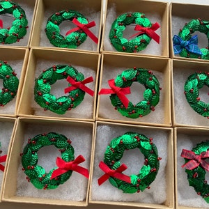 Op de afbeelding: Twaalf kleine groene kransen met rode bessen en rode strikken worden gepresenteerd in individuele dozen. De kransen zijn gemaakt van pailletten en zijn gerangschikt in rijen van vier.