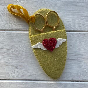 May include: A green felt scissor holder with a red heart and white wings. The holder has a yellow ribbon attached.