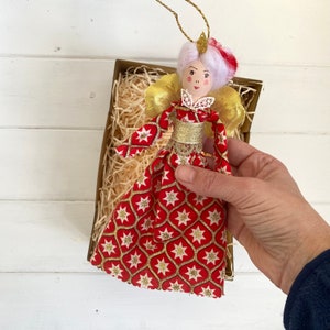 May include: A handmade Christmas ornament featuring a doll with a white wig, red and gold dress, and gold wings. The doll is holding a small white flower.