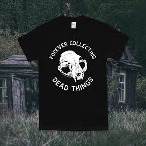 May include: Black t-shirt with a white cat skull graphic and the text "FOREVER COLLECTING DEAD THINGS". The design is centered on the front of the shirt. The shirt is made of a soft material.