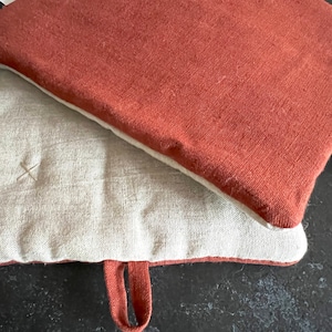 Op de afbeelding: Twee ovenwanten, een is lichtbruin en de andere is roestkleurig. Beide ovenwanten hebben een lusvormige handgreep.