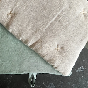 Op de afbeelding: Twee omkeerbare ovenwanten, een is licht beige van kleur en de andere is lichtgroen van kleur. Beide ovenwanten hebben een gestikt X in het midden.