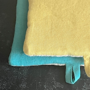 Può includere: Due presine, una blu turchese e una gialla, con un bordo bianco. Entrambe le presine hanno un'ansa a forma di anello.
