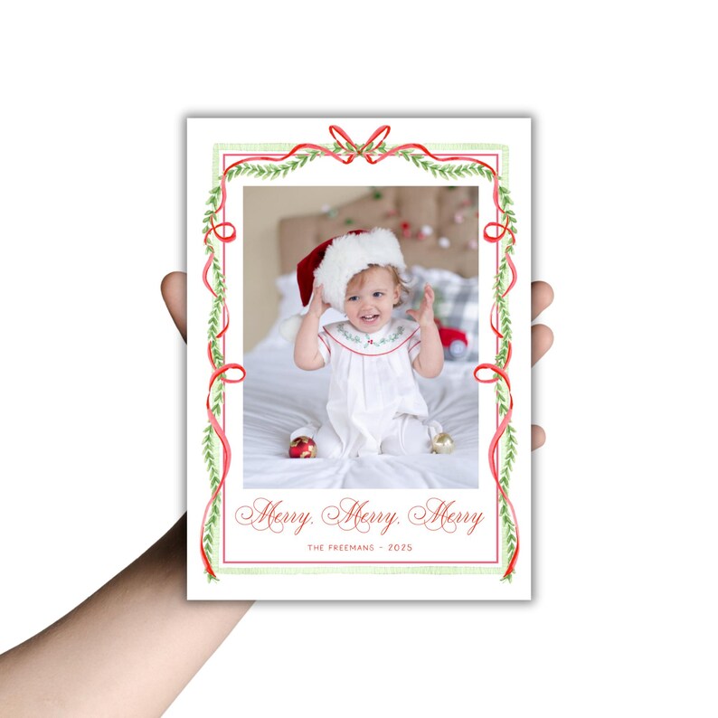 May include: A Christmas card featuring a photo of a baby wearing a Santa hat and a white dress. The card has a red and green watercolor border with the words "Merry, Merry, Merry" and "The Freemans - 2025" printed on it.