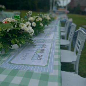 May include: A green and white checkered tablecloth with blue and white train-themed placemats. White chairs are set around the table, which is decorated with white flowers.