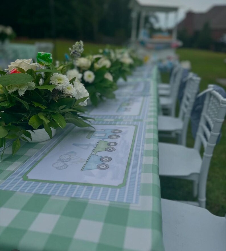 May include: A green and white checkered tablecloth with blue and white train-themed placemats. White chairs are set around the table, which is decorated with white flowers.