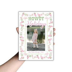 May include: A holiday card featuring a young girl in a pink dress, framed by a border of pink cowboy boots and floral accents. The words "HOWDY Holidays" are displayed in green and pink, with "The Carter Family - 2025" at the bottom.