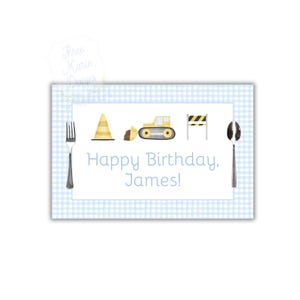 May include: A blue and white checkered placemat with a construction theme. It features a yellow bulldozer, a traffic cone, and a construction sign. The text "Happy Birthday, James!" is printed on the placemat.
