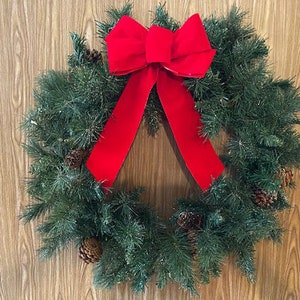 Könnte beinhalten: Ein grüner Weihnachtskranz mit einer großen roten Schleife aus Samt. Der Kranz ist mit Tannenzapfen verziert und hat einen mattierten Look.