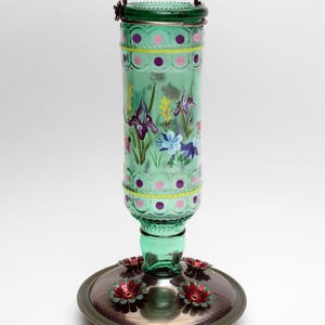 May include: A green glass hummingbird feeder with a cylindrical bottle featuring hand-painted purple and blue floral designs, yellow stripes, and pink polka dots. It has a metal hanging handle and a circular base with red flower-shaped feeding ports.