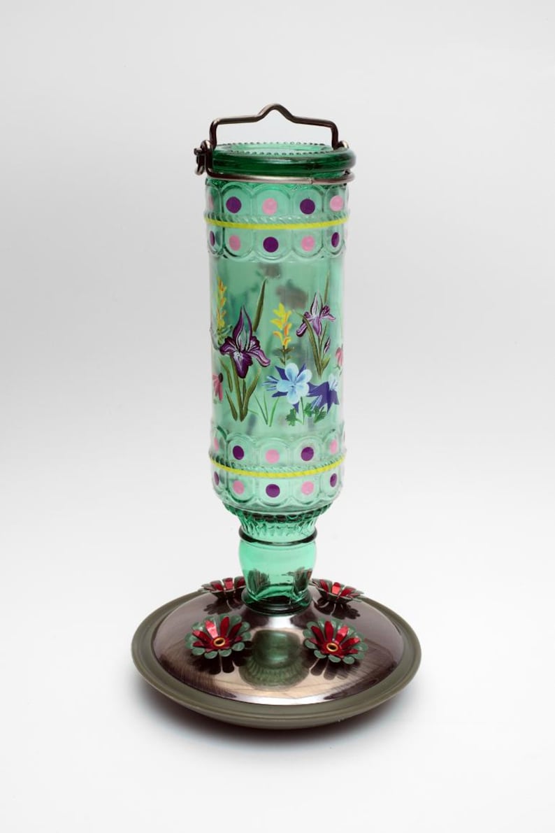 May include: A green glass hummingbird feeder with a cylindrical bottle featuring hand-painted purple and blue floral designs, yellow stripes, and pink polka dots. It has a metal hanging handle and a circular base with red flower-shaped feeding ports.