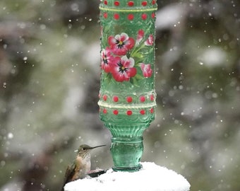 Può includere: Un alimentatore per colibrì con una base in vetro verde e un design floreale rosso e bianco. L'alimentatore è ricoperto di neve e un colibrì è appollaiato sul bordo.
