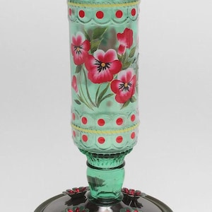 May include: A green glass hummingbird feeder with a cylindrical reservoir featuring hand-painted red and white pansy flowers, yellow lines, and red polka dots. It has a metal wire handle at the top and a circular base with red flower-shaped feeding ports.