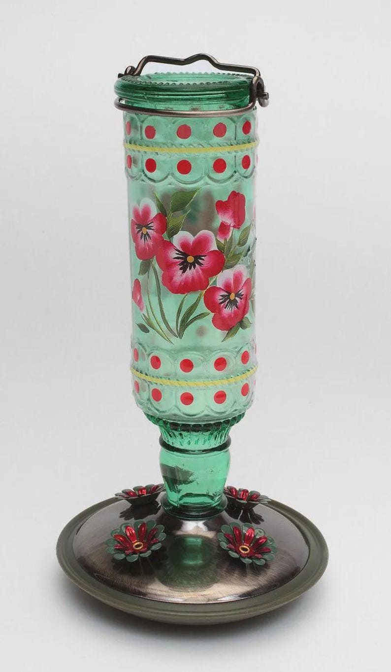 May include: A green glass hummingbird feeder with a cylindrical reservoir featuring hand-painted red and white pansy flowers, yellow lines, and red polka dots. It has a metal wire handle at the top and a circular base with red flower-shaped feeding ports.