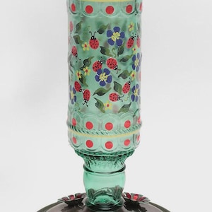 May include: A cylindrical green glass hummingbird feeder featuring a hand-painted pattern of red ladybugs, blue flowers, and green leaves. The top has a metal handle and the base is a circular metallic tray with red flower-shaped feeding ports.