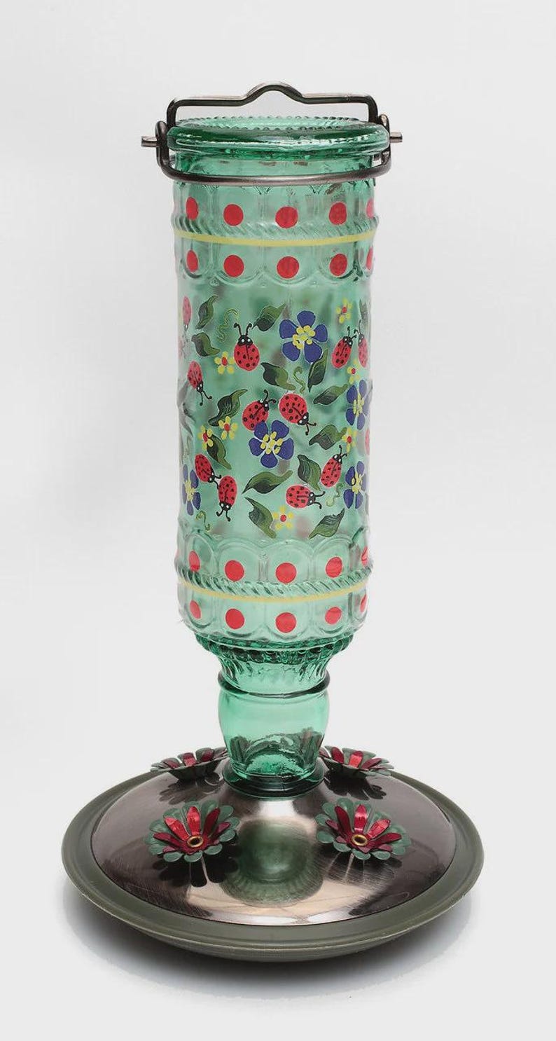 May include: A cylindrical green glass hummingbird feeder featuring a hand-painted pattern of red ladybugs, blue flowers, and green leaves. The top has a metal handle and the base is a circular metallic tray with red flower-shaped feeding ports.