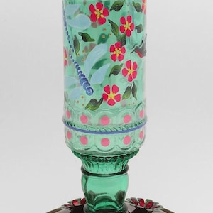 May include: A green glass hummingbird feeder with a cylindrical reservoir featuring hand-painted red flowers, green leaves, and a blue dragonfly. The base is a metallic copper-toned disc with three red and green flower-shaped feeding ports.
