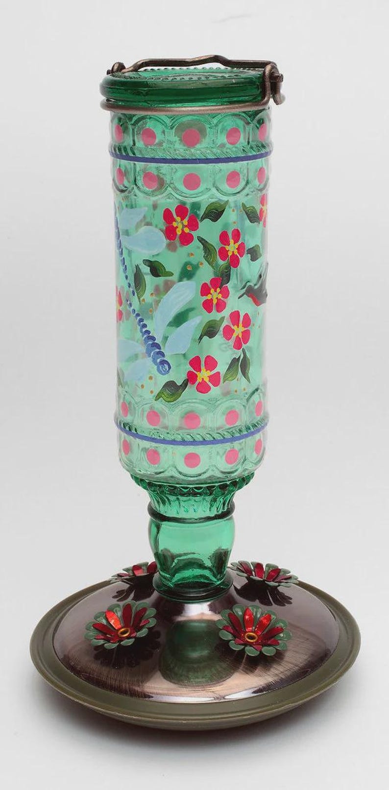 May include: A green glass hummingbird feeder with a cylindrical reservoir featuring hand-painted red flowers, green leaves, and a blue dragonfly. The base is a metallic copper-toned disc with three red and green flower-shaped feeding ports.