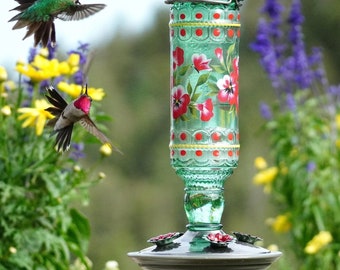 May include: A green glass hummingbird feeder with a floral design. The feeder has a round base and a long, cylindrical body. The feeder is decorated with red, white, and yellow flowers. The feeder is hanging from a hook.