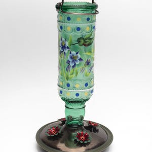 May include: A green glass hummingbird feeder with a cylindrical bottle featuring painted blue and yellow dots, purple columbine flowers, and a small bird. The base is a dark metallic circular tray with several red flower-shaped feeding ports.