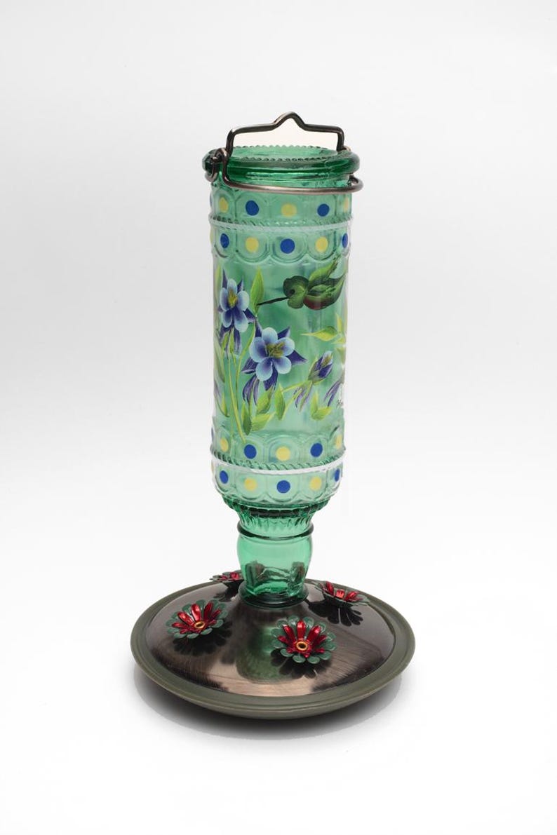 May include: A green glass hummingbird feeder with a cylindrical bottle featuring painted blue and yellow dots, purple columbine flowers, and a small bird. The base is a dark metallic circular tray with several red flower-shaped feeding ports.