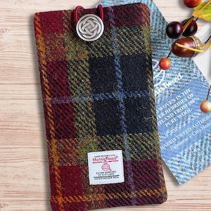 May include: A rectangular Harris Tweed pouch featuring a multi-colored plaid pattern in shades of burgundy, olive green, and black. The pouch is secured with a red elastic loop and a silver-toned metal button with a Celtic knot design.