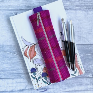 Peut inclure: Une trousse magenta avec une fermeture éclair argentée et une languette violette, posée sur un carnet avec une illustration de vache des Highlands. Deux stylos argentés et noirs sont également présents. La trousse a un motif à carreaux.