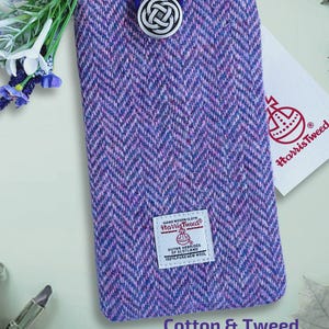Puede incluir: Una funda rectangular para teléfono de Harris Tweed con un patrón de espiga en tonos morado y azul. Presenta un botón circular plateado con un nudo celta y un cierre de bucle elástico azul. Una etiqueta blanca de Harris Tweed está cosida.