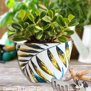 Peut inclure: Un pot en tissu blanc avec un motif de feuilles tropicales dans des tons de vert, bleu et jaune. Le pot est rempli d'une plante verte et est posé sur une surface en bois.
