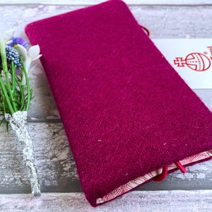 Pu&ograve; includere: Una copertina rettangolare in lana magenta con trama testurizzata, dotata di una chiusura elastica rossa e di un'etichetta bianca con il logo Harris Tweed. Un piccolo bouquet di fiori artificiali viola e bianchi &egrave; posizionato accanto.