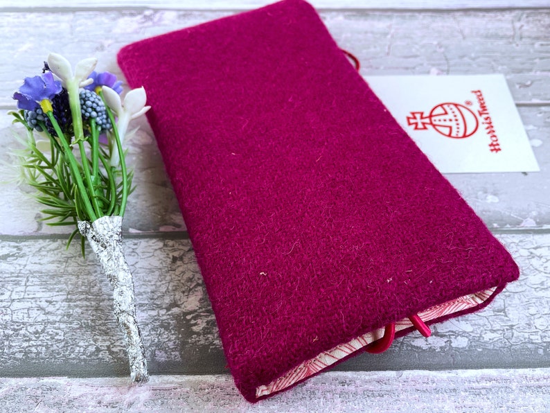 Pu&ograve; includere: Una copertina rettangolare in lana magenta con trama testurizzata, dotata di una chiusura elastica rossa e di un'etichetta bianca con il logo Harris Tweed. Un piccolo bouquet di fiori artificiali viola e bianchi &egrave; posizionato accanto.