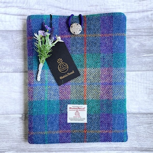 Puede incluir: Una funda para libros Harris Tweed a cuadros azules, turquesas y morados. Cuenta con un botón plateado decorativo y un pequeño arreglo floral. Se adjunta una etiqueta negra con texto dorado. La funda tiene un acabado texturizado.